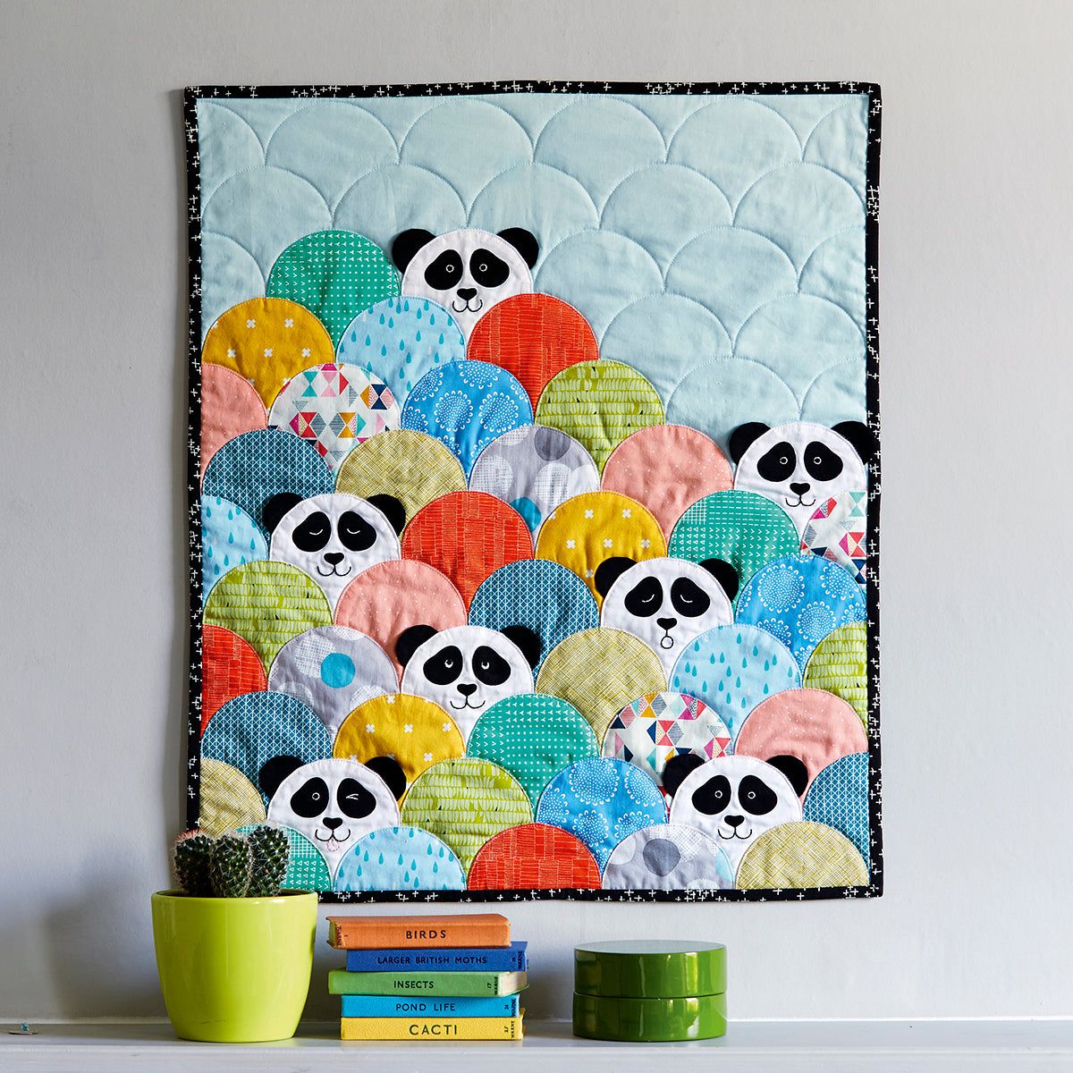 A colorful quilt featuring panda faces in a pattern with scallop edges, displayed on a wall with potted plants and books below.