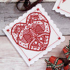Cross stitch red design on a white fabric square with Christmas decorations in the background.