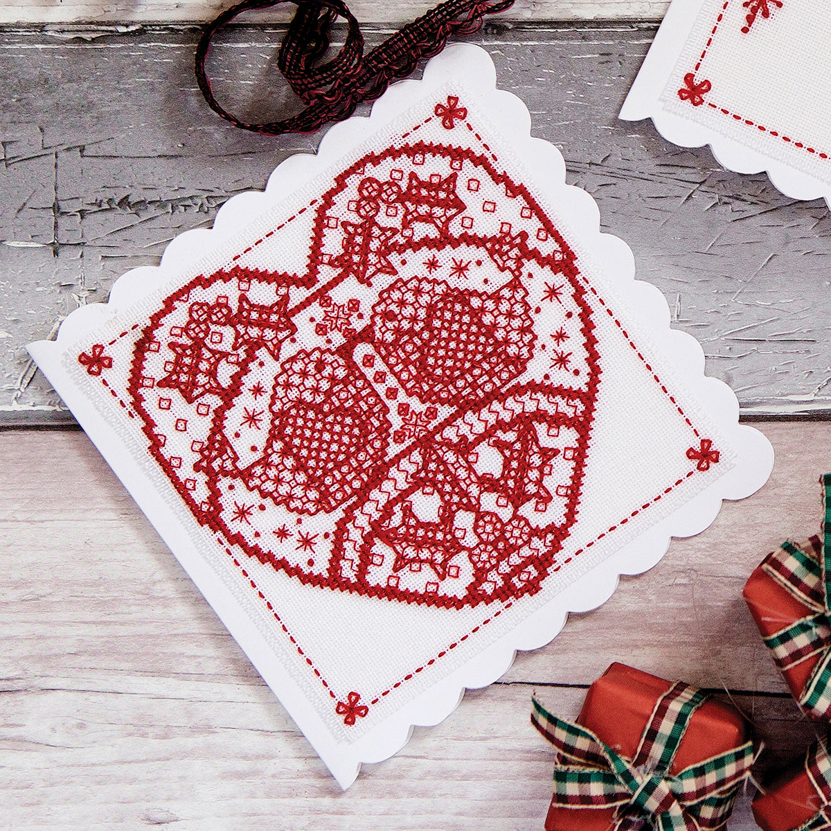 Cross stitch red design on a white fabric square with Christmas decorations in the background.