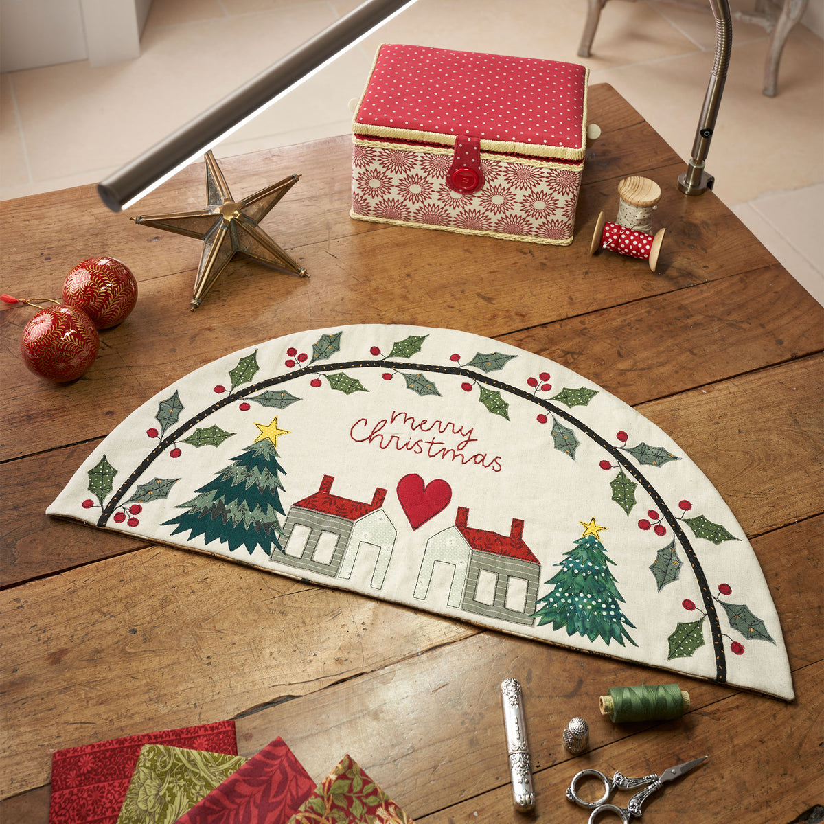 Half-circle Christmas wall hanging with holly, houses, and trees, says "Merry Christmas." On a wooden table with a red polka-dot box and sewing supplies. Festive.